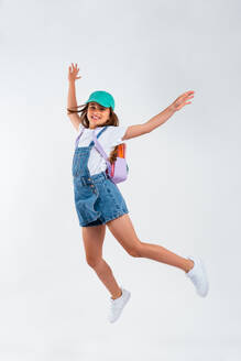 Full body of happy energetic teen girl in denim jumpsuit jumping and raising arms while dancing against white background with open eyes - ADSF45587