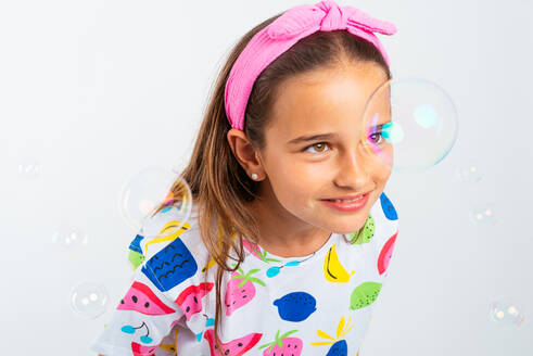 Happy little girl wearing colorful clothes and headband looking at soap bubbles while smiling against white background in studio shot - ADSF45594