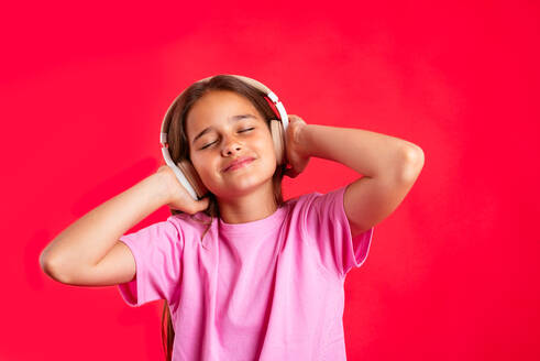 Cheerful girl listening to music in headphones with hands on head and elbow up while standing against red background with closed eyes - ADSF45596