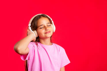 Cheerful girl listening to music with headphones holding the headphones in her hand standing against a red background and with her eyes clenched - ADSF45597