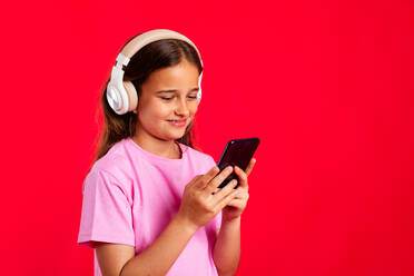 Happy cute girl in purple casual clothes smiling and listening to music in wireless headphones while browsing smartphone against red background - ADSF45598