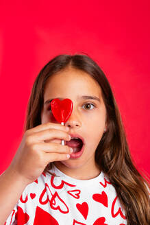 Portrait of surprised girl in casual clothes with red heart shaped lollipop looking at camera with omg bewildered open mouth while standing - ADSF45599