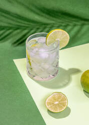 From above of transparent crystal glass of cocktail drink with lime slice and ice placed on white sheet on green surface near shade of leaves - ADSF45603