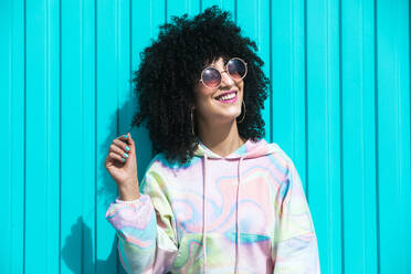 Positive young Hispanic female with Afro hairstyle and sunglasses smiling and touching hair against blue wall in sunlight - ADSF45654