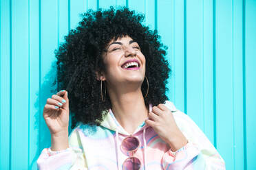 Happy young Hispanic female with Afro hairstyle in sportswear and sunglasses with closed eyes laughing against blue background - ADSF45655