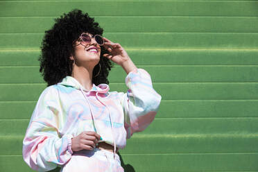 Happy young Hispanic female with Afro hairstyle and closed eyes adjusting sunglasses with hand against green wall in sunlight - ADSF45656