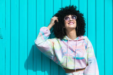 Happy young Hispanic female with Afro hairstyle and closed eyes touching hair with hand against blue wall in sunlight - ADSF45657