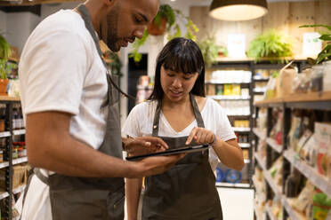 Young African American male and female sales staff in apron standing in between cupboards with grocery items and looking at screen of tablet while browsing - ADSF45659