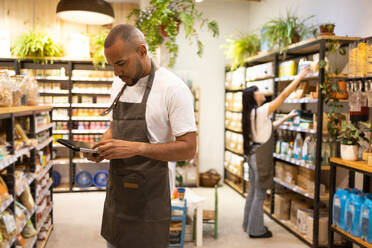 Side view of African American male seller in apron standing near shelves with groceries and looking at screen of tablet while checking information - ADSF45661