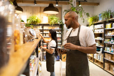 Side view of African American African American male seller in apron standing near shelves with groceries and to taking notes on his tablet - ADSF45662