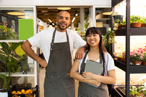 Positive young African American sales staff in apron smiling and looking at camera while male standing with hand on shoulder of female with tablet - ADSF45666