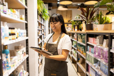Happy young Asian female seller in apron smiling and browsing tablet while standing near shelves with various goods during work in modern store - ADSF45669