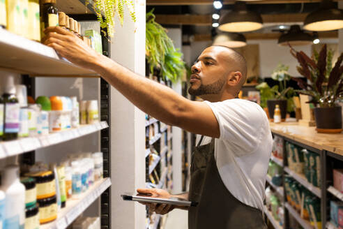 Side view of concentrated young African American male seller in apron taking product from shelf while standing with tablet during work process in cosmetic shop - ADSF45670