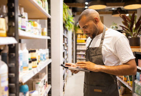 Side view of focused young African American male seller in apron standing near shelves with various goods and checking information in tablet while working - ADSF45671