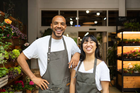 Cheerful young multiracial colleagues in aprons smiling and looking at camera while standing together in floristry store during work process - ADSF45672