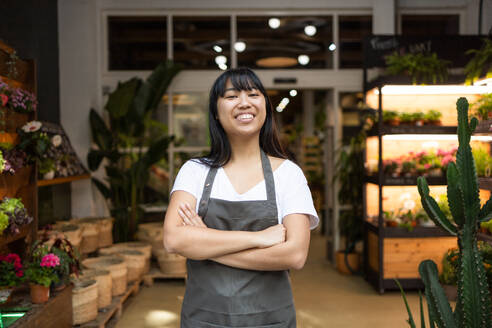 Happy young Asian florist in apron smiling and looking at the camera while standing with her arms crossed while working in a florist shop - ADSF45674