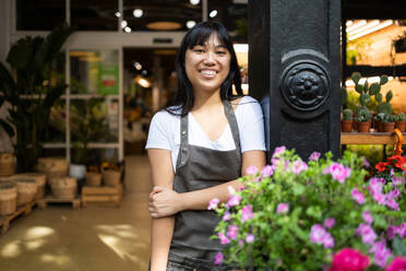 Happy young Asian female florist in apron smiling and looking at camera while standing near counter with blooming flowers during work in floristry shop - ADSF45675