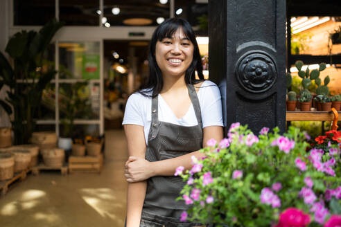 Happy young Asian female florist in apron smiling and looking at camera while standing near counter with blooming flowers during work in floristry shop - ADSF45675