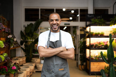 Cheerful young African American male florist in apron smiling and looking at camera while standing near blooming flowers during work day - ADSF45677