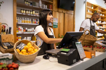 Cheerful Asian female seller in apron smiling and using computer while standing at counter with her colleague and various products while working in a supermarket - ADSF45678