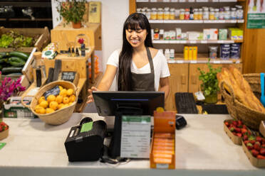 Happy young Asian female seller in apron smiling and using computer while standing at counter with various products during work in supermarket - ADSF45680