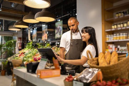 Happy young multiracial coworkers in aprons smiling and using monitor to enter information while standing at counter together during work - ADSF45681