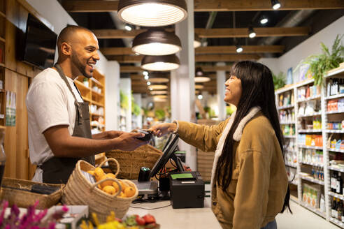 Happy customer smiling at the worker while paying with contactless credit card at a shopping terminal looking at each other - ADSF45684