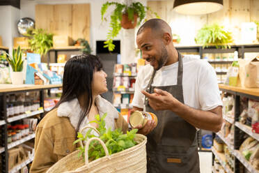 Positive young Asian female customer with basket of vegetables smiling and speaking to African American male seller in apron in modern supermarket - ADSF45685