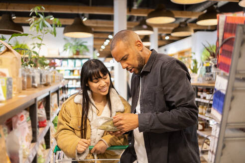 Concentrated young multiethnic couple in casual wear choosing goods in supermarket together while shopping with grocery cart during sale - ADSF45686