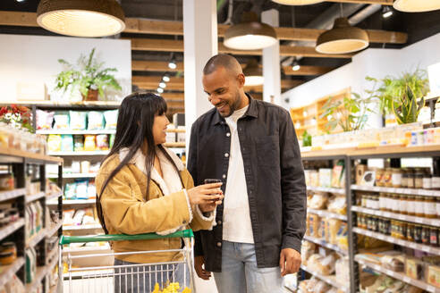 Cheerful young multiethnic couple in casual wear choosing goods in supermarket together while shopping with grocery cart during sale - ADSF45687