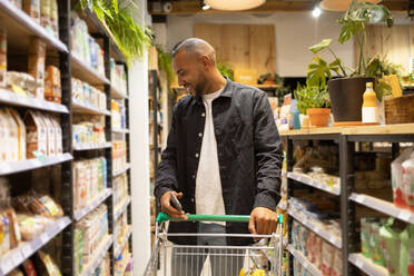 Positive young African American male customer in casual outfit with shopping cart smiling and choosing food from shelves - ADSF45691