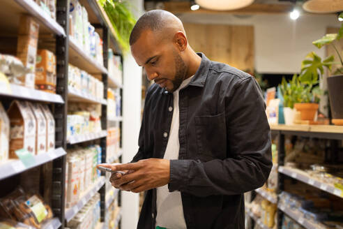 Focused young African American male customer in casual clothes messaging on mobile phone while standing near shelves in grocery shop - ADSF45694