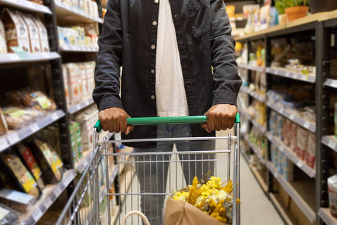 Cropped anonymous man in casual outfit with shopping trolley picking food from shelves while in supermarket shopping - ADSF45695