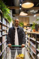 Positive young African American man in casual outfit with shopping trolley smiling and looking at the camera while in a supermarket - ADSF45696