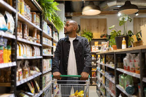 Positive young African American man in casual outfit with shopping trolley looking away while in a supermarket - ADSF45697