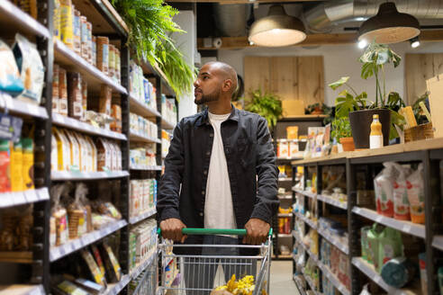 Positive young African American man in casual outfit with shopping trolley looking away while in a supermarket - ADSF45697