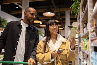 Low angle of cheerful multiethnic couple in casual clothes with shopping trolley and smartphone choosing goods while buying products together - ADSF45699