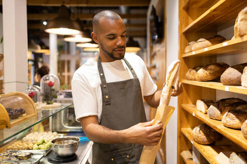Positive young African American male seller in apron putting fresh baked bread into paper bag while standing near shelves during work process - ADSF45700