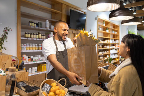 Positive young African American male seller in apron smiling and giving paper bag with products to Asian female customer in modern supermarket - ADSF45702