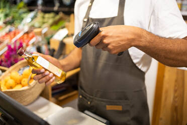 Unrecognizable male seller in apron standing near counter and scanning barcode on oil bottle with handheld wireless scanner while working at shop - ADSF45703