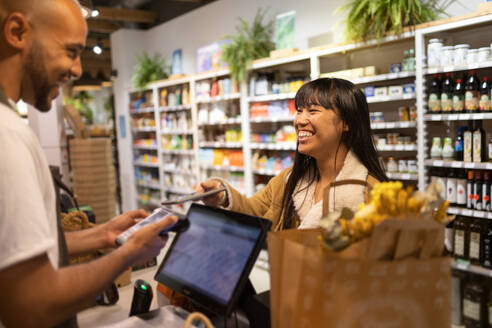 Salesman African American male in apron a counter with a POS terminal while an Asian customer pays electronically for purchases made with a smartphone - ADSF45705