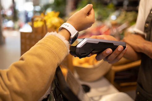 Side view of anonymous female in warm clothes making contactless electronic payment with smartwatch on POS terminal held by unrecognizable salesman - ADSF45707