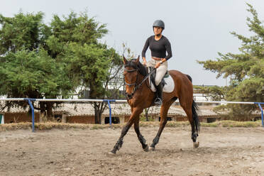 Front view of young female equestrian in helmet and riding boots riding chestnut horse on huddle race in countryside under cloudy sky - ADSF45708