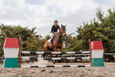 From below of young female equestrian in helmet and riding boots riding chestnut horse on huddle race over barrier in countryside under cloudy sky - ADSF45709