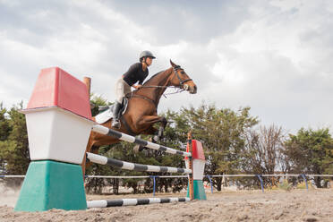 Side view of young female equestrian in helmet and riding boots riding chestnut horse on huddle race over barrier in countryside under cloudy sky - ADSF45710