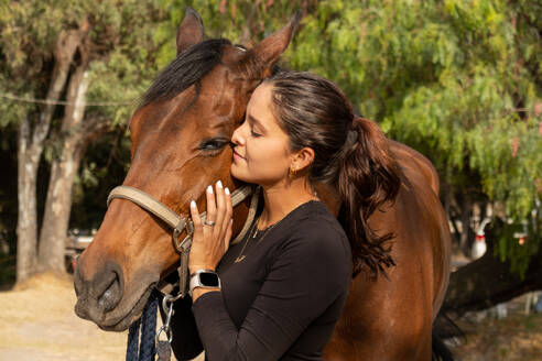 Side view of young happy female in casual clothes embracing chestnut horse while standing and smiling on ranch in daylight - ADSF45711