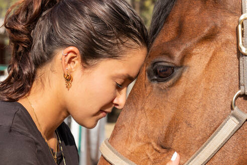 Side view of young female in casual clothes embracing chestnut horse while standing and smiling on ranch in daylight - ADSF45712