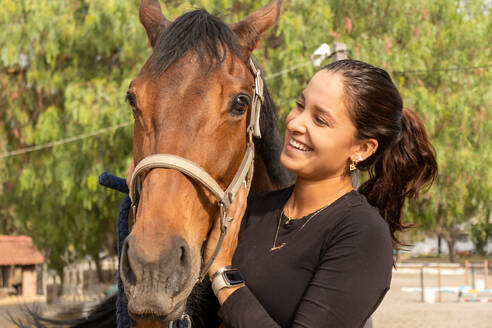 Front view of young happy female in casual clothes embracing chestnut horse while standing and smiling on ranch in daylight - ADSF45713