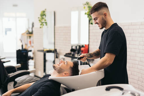 Side view of young barber washing closed eyes man's hair in barber shop against burred background - ADSF45714