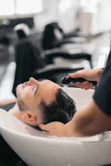 Cropped anonymous young barber washing man's hair against blurred background in barber shop - ADSF45715